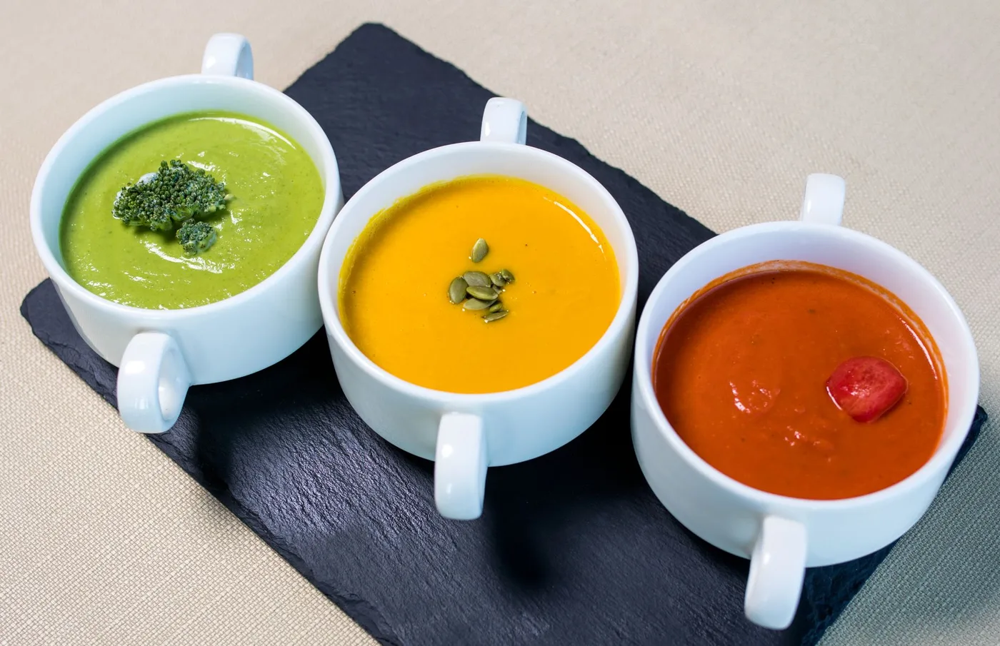 A variety of vegetarian soups presented in white mugs, featuring broccoli, pumpkin, and tomato flavors.