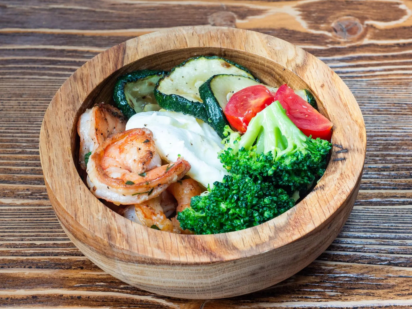A wooden bowl filled with shrimps, broccoli, zucchini, and tomatoes on a rustic table.