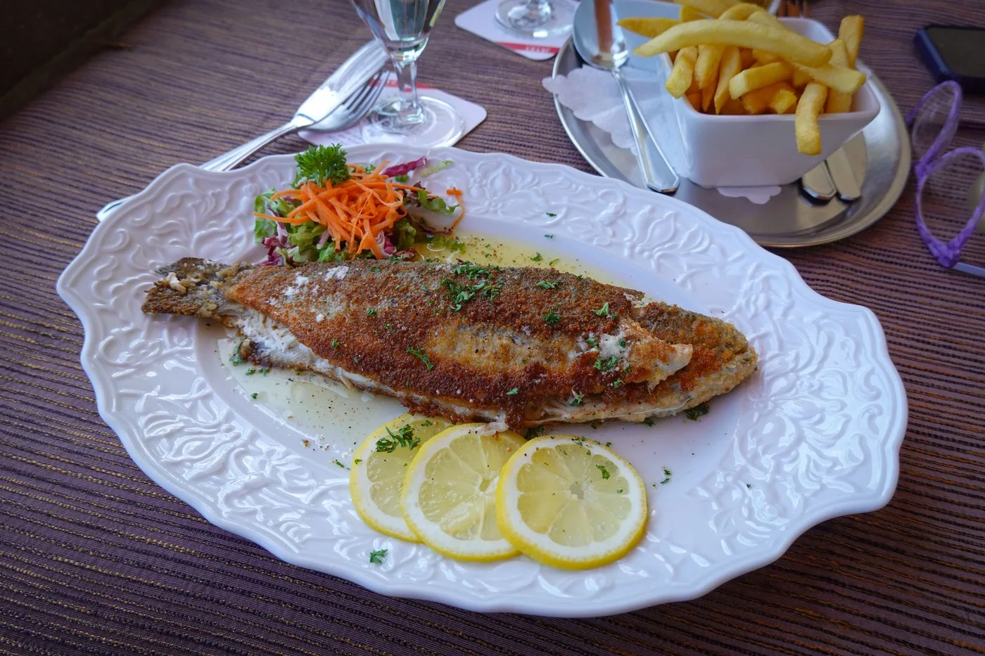 Grilled fish served with lemon slices, fries, and a side salad on a decorative plate.
