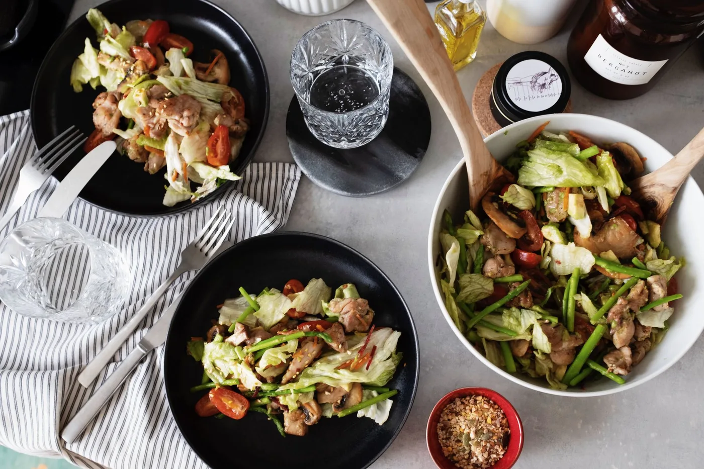 Top view of fresh salad served on black plates with forks and knifes from big white bowl on table with glasses of water