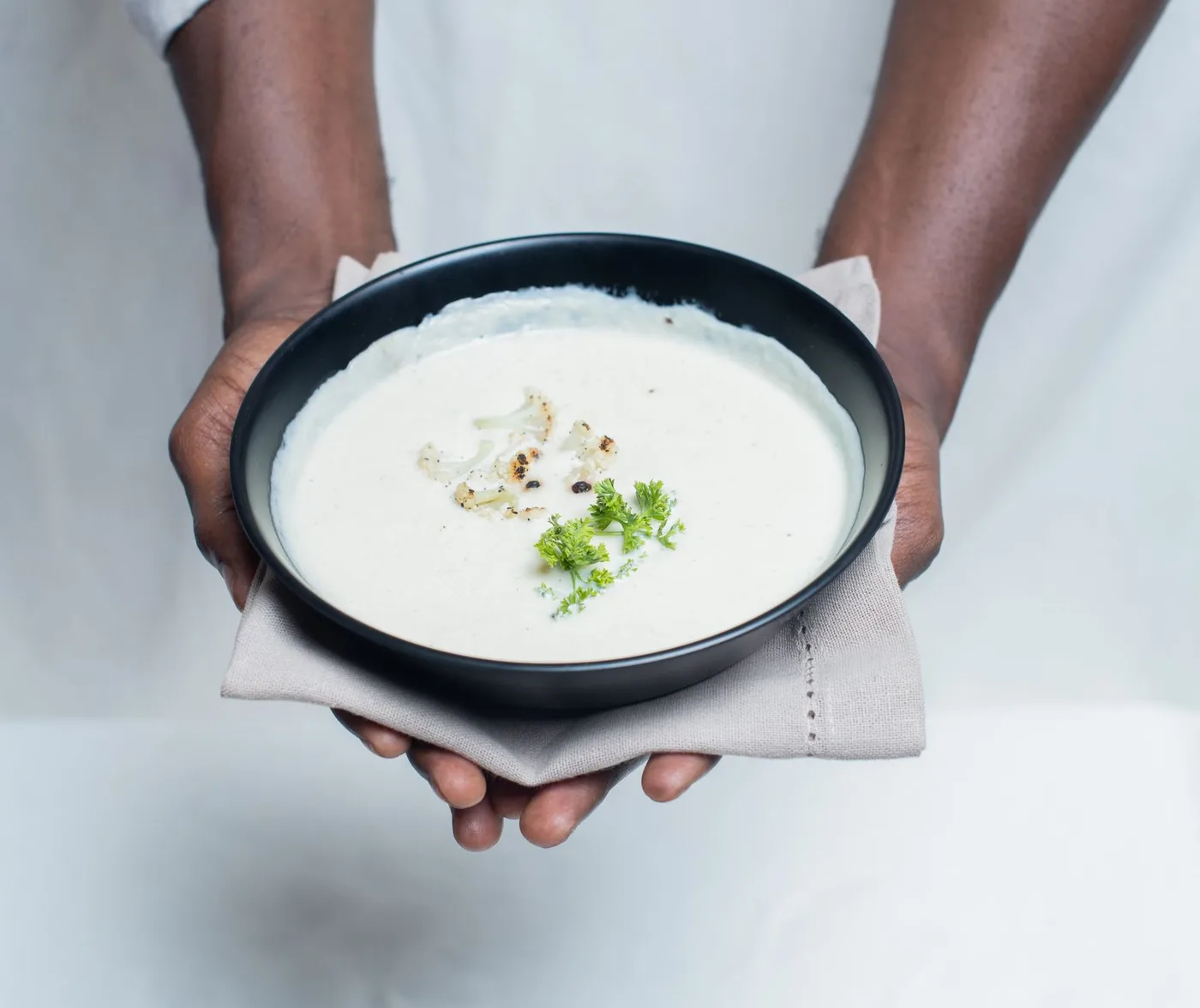 A close-up of hands holding a bowl of creamy cauliflower soup garnished with parsley.